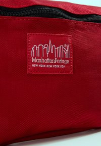 Red nylon pouch with a zip closure features a white Manhattan Portage logo, depicting a skyline and brand name. Textured fabric detail visible.