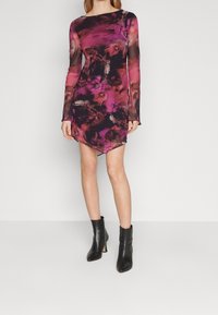 Robe moulante à manches longues de couleur noire avec un motif floral rose et violet, dotée d'un ourlet asymétrique. Associée à des bottines noires à la cheville.