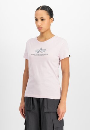Mujer con cabello trenzado lleva una camiseta rosa claro de Alpha Industries y pantalones cargo negros sobre un fondo blanco liso.