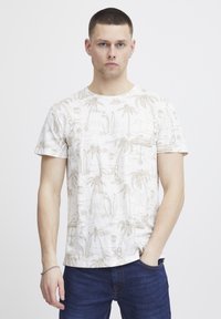 Camiseta de manga corta color crema con un patrón de palmera y tabla de surf en beige. Cuello redondo y textura suave, combinada con jeans azules.