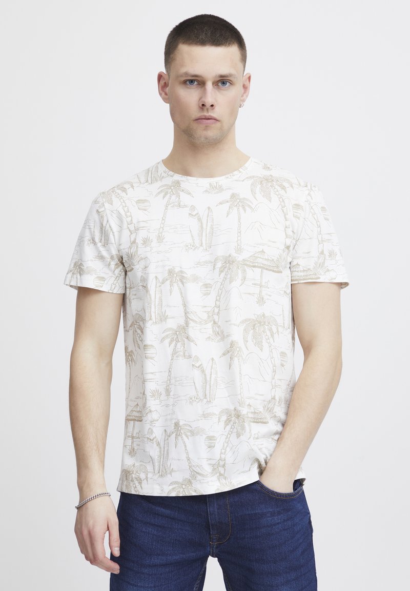 Camiseta de manga corta color crema con un patrón de palmera y tabla de surf en beige. Cuello redondo y textura suave, combinada con jeans azules.