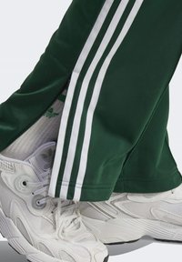 Vihreät verkkarit, joissa on kolme valkoista raitaa sivuilla ja vetoketju lahkeen resorissa. Yhdistetty valkoisiin urheilukenkiin, joissa on tekstuurilliset yksityiskohdat.