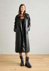 Manteau trench en cuir synthétique noir avec un design à double boutonnage, grands revers et poches latérales, associé à des bottines noires à lacets.