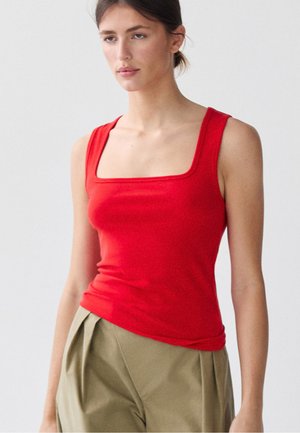 Femme portant un haut rouge sans manches avec un col carré et un pantalon beige taille haute, debout devant un fond clair uni.