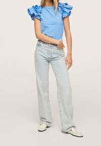 Haut bleu clair avec manches à volants, associé à un jean taille haute à lavage clair et des chaussures blanches à enfiler. Textures lisses visibles.