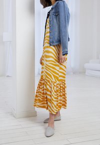 Abito maxi con strisce gialle e bianche, orlo svasato e dettagli arricciati, abbinato a una giacca di denim chiaro e a delle scarpe slip-on grigie.