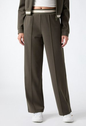 Femme portant un pantalon large couleur vert olive avec une ceinture à logo, des baskets blanches et une veste courte assortie.