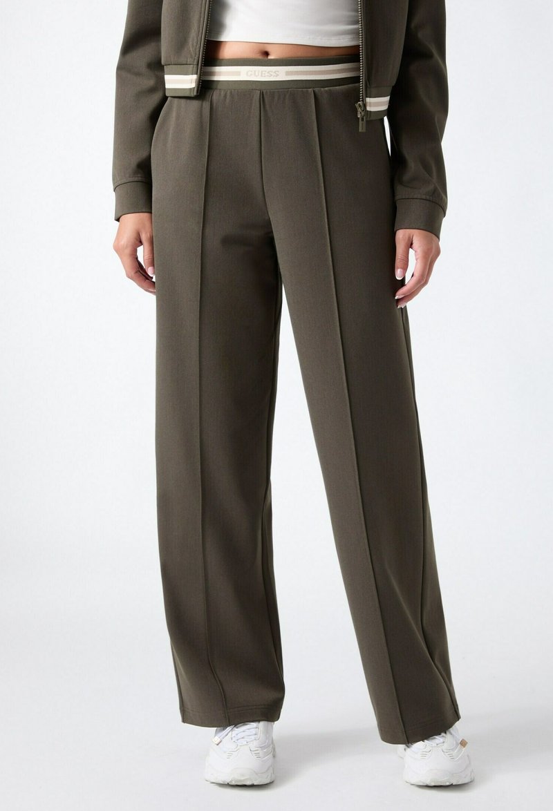 Femme portant un pantalon large couleur vert olive avec une ceinture à logo, des baskets blanches et une veste courte assortie.