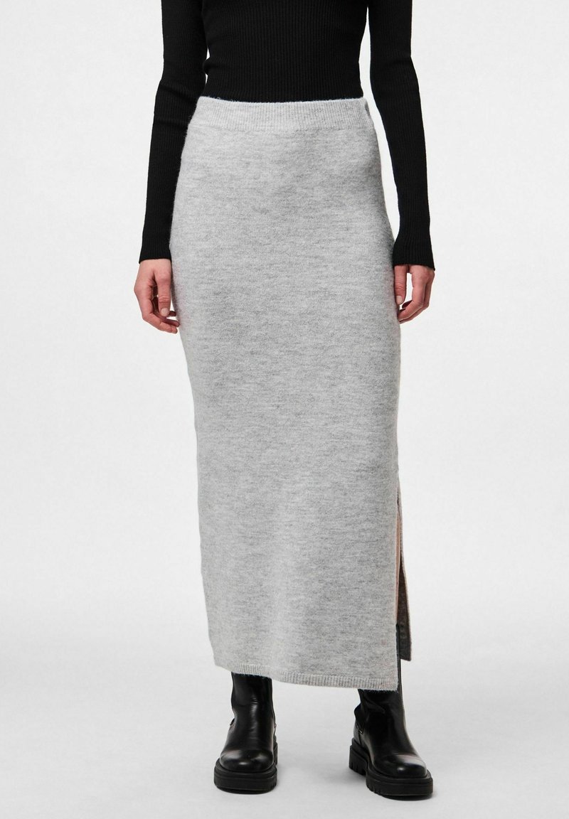 Femme portant une jupe longue en maille gris clair avec une fente latérale, un haut à manches longues noir et des bottines noires épaisses, debout devant un fond blanc.