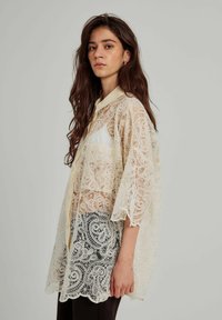 Camicia in pizzo trasparente in stile kimono color crema con motivi intricati, maniche corte a sbuffo e chiusura con bottoni. Indossata sopra pantaloni scuri.