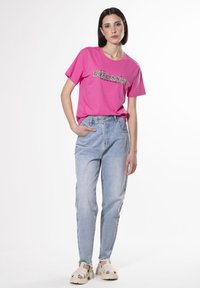Camiseta de algodón rosa con gráfico de "#Jefe", combinada con vaqueros de mezclilla azul claro. La modelo lleva sandalias beige con suela gruesa.