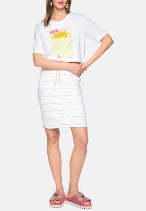 Frau trägt ein weißes T-Shirt mit Grafik, einen texturierten Rock mit pastellfarbenen Streifen und Zugband sowie pink glitzernde Sandalen mit Schnallen, steht mit verschränkten Armen.