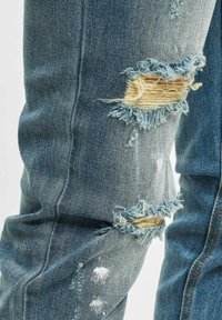 Blå denim jeans med slitte rifter på knærne med frynsete kanter. Klassisk fem-lomme design og synlige sømdetaljer.