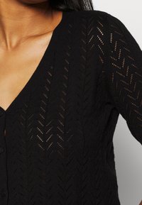Cárdigan de punto negro con cuello en V, cierre de botones y un patrón de espiga con detalles de malla transparente en todo el diseño.