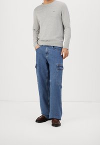 Grauer Rundhals-Pullover kombiniert mit lockeren blauen Cargo-Jeans mit Seitentaschen. Braune Schuhe vervollständigen das Outfit.