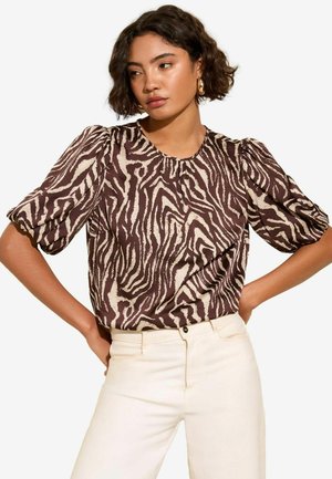 Blusa a strisce zebra marrone e crema con maniche corte a palloncino e scollatura tonda. Presenta una superficie liscia e una vestibilità ampia. Abbinata a pantaloni chiari.