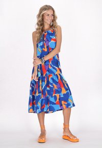 Farbenfrohes, ärmelloses Kleid in Blau mit abstrakten Mustern in Orange und Lila. Verfügt über einen gerafften Ausschnitt und einen gestuften Saum. Kombiniert mit orangenen Sandalen.