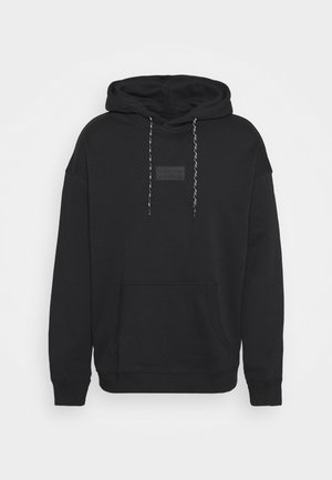Sweat à capuche noir avec une poche avant et des cordons de serrage gris, porté avec un pantalon noir et des baskets blanches avec des rayures noires et des accents marron.