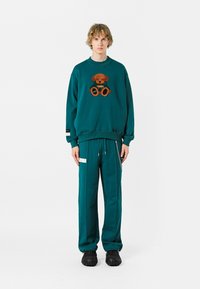Sudadera color teal con bordado de oso de peluche, combinada con joggers a juego que tienen detalles con cremallera y bolsillos laterales. El material parece suave y acogedor.
