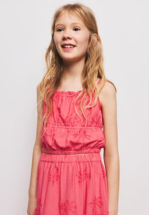 Lachend jong meisje met lang blond haar, gekleed in een mouwloze roze jurk met subtiele bloemenpatronen tegen een lichte achtergrond.