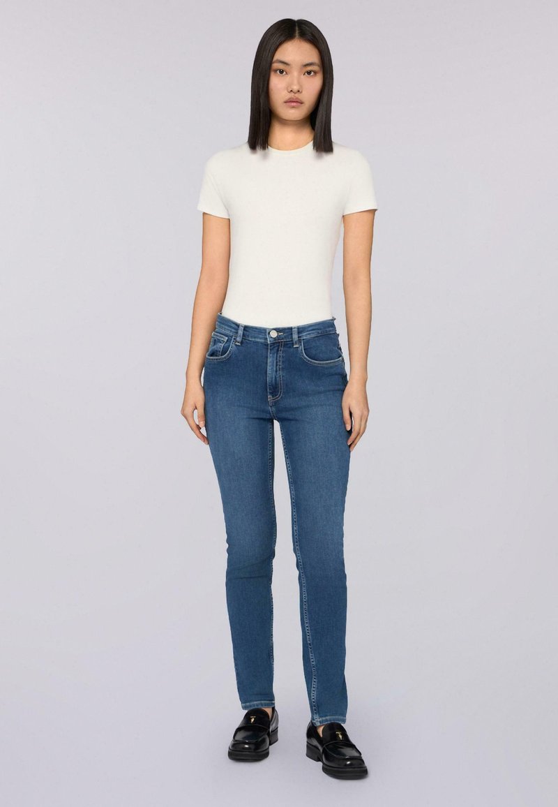 Jeans de mezclilla azul ajustados con cinco bolsillos, cierre de botón frontal y una textura ligeramente desgastada, combinados con una camiseta blanca entallada.