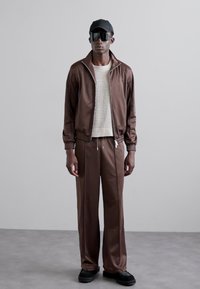 Brun satin treningsdrakt med glidelåsjakke og bukser med snor, kombinert med en teksturert kremfarget strikkegenser og svarte tilbehør.
