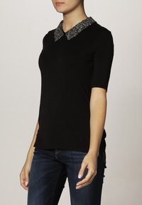 Kvinna klädd i en svart kortärmad topp med en glittrande dekorerad krage och mörkblå jeans, stående mot en enfärgad bakgrund.