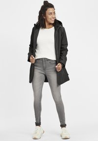 Chaqueta impermeable negra con capucha, llevada sobre un suéter crema. Jeans grises, zapatillas blancas y amarillas completan el atuendo. Estilo minimalista.