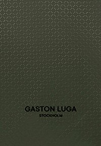 Mörkgrön texturerad yta med en diamantmönster, där varumärket "GASTON LUGA" och "STOCKHOLM" är tryckt i svart.