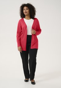 Cardigan rouge en matériau doux, design à l'avant ouvert, associé à un haut blanc et un pantalon noir, chaussures plates noires, cheveux bouclés texturés.