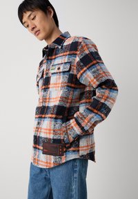 Camicia in flanella con un motivo a quadri arancione, blu e nero. Presenta due tasche sul petto e un'etichetta con il marchio nella parte anteriore in basso.