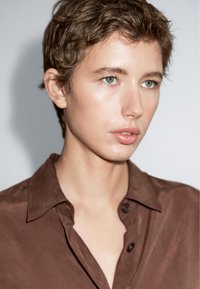 Brun skjorte med knapper og en krave, blød tekstur og let glans. Modellen har kort, rodet hår og blå øjne, neutral baggrund.