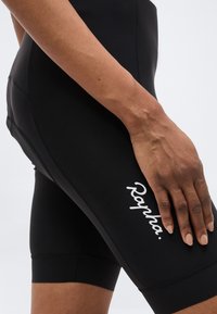 Gros plan sur la main d'une personne reposant sur un short de cyclisme Rapha noir, sur un fond clair uni.