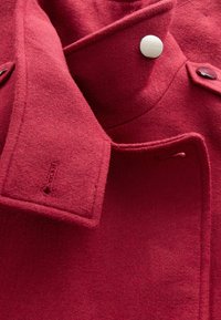 Red wool coat with a structured collar, featuring a white button and subtle stitching details. Soft texture, tailored design, and minimal accents.
