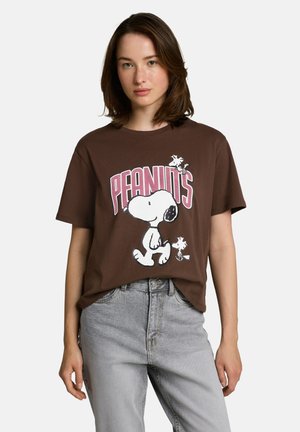 Młoda kobieta w brązowym T-shircie Peanuts z postaciami Snoopy i Woodstock, w połączeniu z jasnoszarymi jeansami na jednolitym tle.