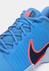 Κοντινό πλάνο μπλε αθλητικού παπουτσιού με μπλε κορδόνια, μαύρο και πορτοκαλί λογότυπο Nike swoosh, και υφή από δίχτυ και συνθετικό επάνω μέρος.