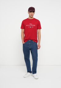 Červené bavlněné tričko s bílým logem "HILFIGER", skombinované s modrými riflovými džíny a bílými teniskami, doplněné tmavou čepicí.