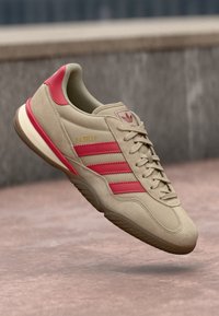 Beige kedas ar sarkaniem svītrām, izgatavoti no zamšādas, ar zemu profilu, noapaļota purngalu un teksturētu zoli. Iezīmē zelta "Gazelle" zīmolu.