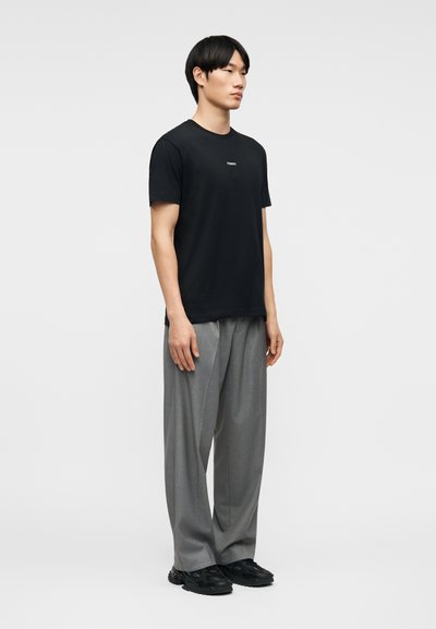 Νέος άνδρας που στέκεται, φοράει μαύρο κοντομάνικο μπλουζάκι, άνετο γκρι παντελόνι και μαύρα παπούτσια μπροστά σε απλό ανοιχτό φόντο.