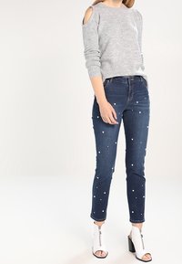 Grijze gebreide trui met uitsnijdingen op de schouders, gecombineerd met blauwe denim jeans versierd met witte kralen, en witte enkellaarsjes met hak.