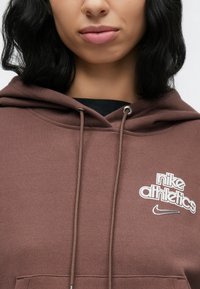 Καφέ φούτερ με κουκούλα από μαλακό ύφασμα, με κορδόνι στην κουκούλα, τσέπη μπροστά και λευκό κεντητό λογότυπο με λεπτομέρεια σβούρας.