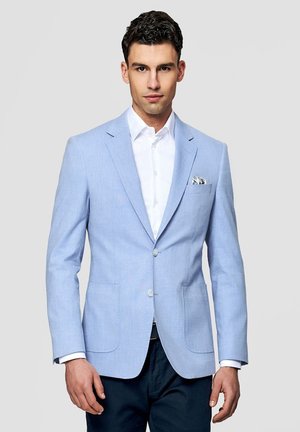 Mężczyzna w jasnoniebieskiej marynarce, białej koszuli, ciemnych spodniach, stojący na tle jednolitego szarego tła, zwrócony przodem.