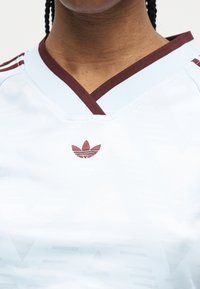 Prim-plan cu o persoană care poartă un tricou sport alb cu margini bordo și un logo bordo central pe piept.