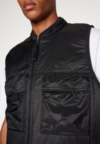 Μαύρο νάιλον γιλέκο με υψηλό γιακά, φερμουάρ μπροστά και δύο μεγάλα τσέπες. Διαθέτει μια διακριτική υφή πλέγματος και κεντητό λογότυπο στο στήθος.