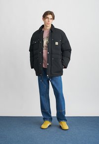 Μαύρο padded μπουφάν με δύο μπροστινές τσέπες, φορεμένο πάνω από ροζ φούτερ και μπλε τζιν, συμπληρωμένο με κίτρινα αθλητικά παπούτσια.