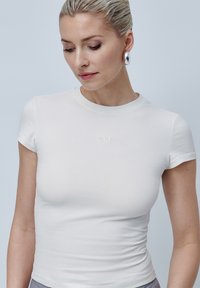 Camiseta blanca ajustada de manga corta, con cuello redondo y un sutil logo bordado en el pecho. Tela de algodón lisa y elástica.