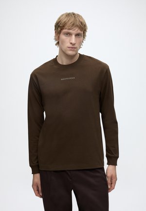 Jonge man met licht haar, gekleed in een bruin shirt met lange mouwen en een donkere broek, staand voor een effen witte achtergrond.