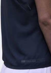 Gros plan d'une personne portant un t-shirt de sport bleu marine avec le mot "TENNIS" imprimé près de l'ourlet et un short blanc visible en dessous.