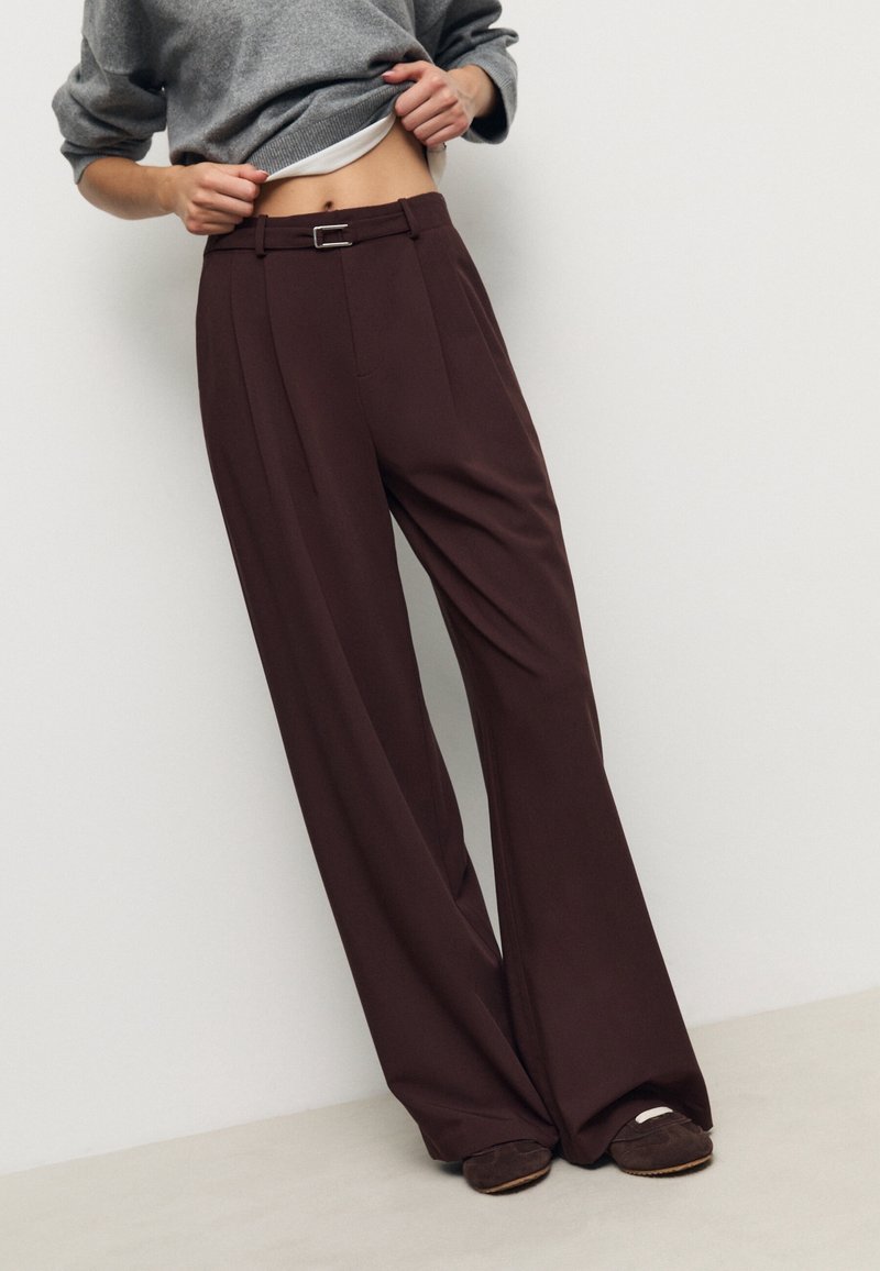 Personne soulevant un pull gris, portant un pantalon large taille haute marron foncé avec ceinture et chaussures foncées sur un fond clair uni.