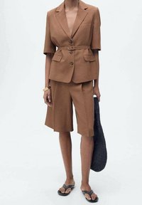 Blazer marron sur mesure à manches courtes, short assorti longueur genou, sac noir tissé, accessoirisé de bracelets dorés et sandales.